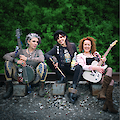 Das Bild zeigt eine Musikgruppe bestehend aus drei Personen, die auf Verstärkern oder Boxen sitzen vor einem grünen Hintergrund aus Pflanzen. Die Musiker halten verschiedene Instrumente - eine Banjo/Banjo-Gitarre links, ein Schlagzeug-Stick und eine Flasche in der Mitte, sowie eine E-Gitarre rechts - und tragen unterschiedliche stilvolle Outfits mit einem rockigen, alternativen Erscheinungsbild.