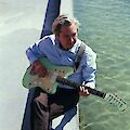 Ein Mann mit hellblondem Haar sitzt auf einer Betonkante am Wasser und spielt eine mintgrüne Fender-Gitarre. Er trägt ein hellblaues Hemd und dunkle Hose, und das türkisfarbene Wasser im Hintergrund verleiht dem Bild eine entspannte, sommerliche Atmosphäre.